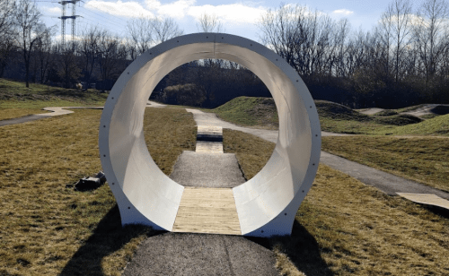A large circular tunnel structure in a grassy park, with a pathway leading through it under a clear blue sky.