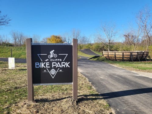 Sign for "Cliffs Bike Park" with a bike icon, surrounded by green grass and a dirt ramp in the background. Clear blue sky.