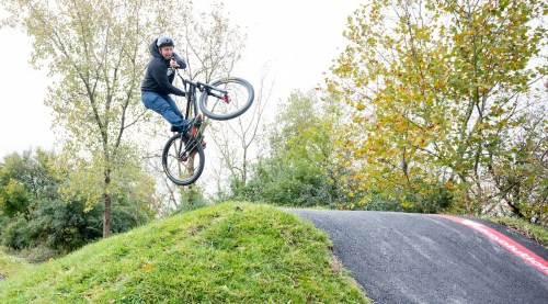 A cyclist performs a jump on a bike over a dirt ramp, surrounded by trees and greenery.