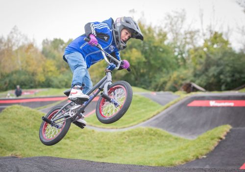 A young boy in a helmet performs a jump on a BMX bike at a pump track, surrounded by greenery.