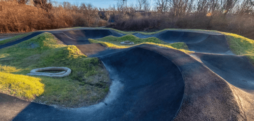 A winding pump track with grassy mounds, surrounded by trees and shrubs, under a clear blue sky.