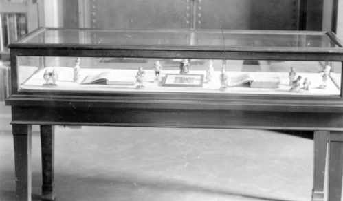 A glass display case containing small figurines and books, set in a room with a blurred background.