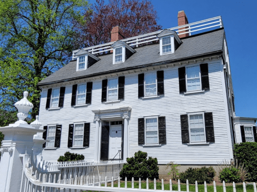 A historic white house with black shutters, a fenced yard, and a clear blue sky in the background.