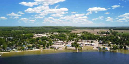 Aerial view of a small town by a lake, surrounded by green trees and fields under a blue sky with fluffy clouds.