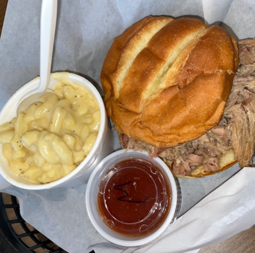 A plate featuring a pulled pork sandwich, creamy macaroni and cheese, and a small cup of barbecue sauce.