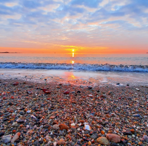 A serene beach at sunset, with colorful clouds reflecting on the water and pebbles scattered along the shore.
