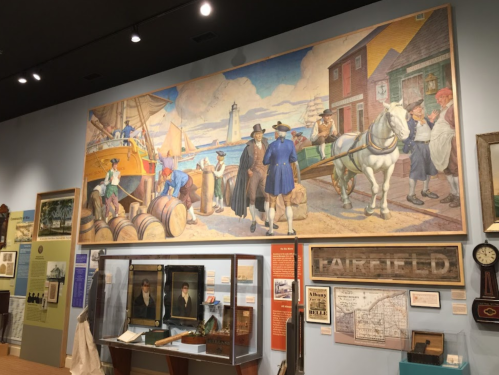 A mural depicting a coastal scene with people, a horse, and a lighthouse, surrounded by historical artifacts in a museum.