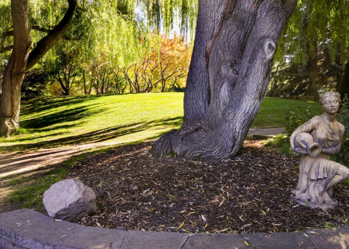 A serene park scene featuring a large tree, a stone statue, and colorful autumn foliage in the background.