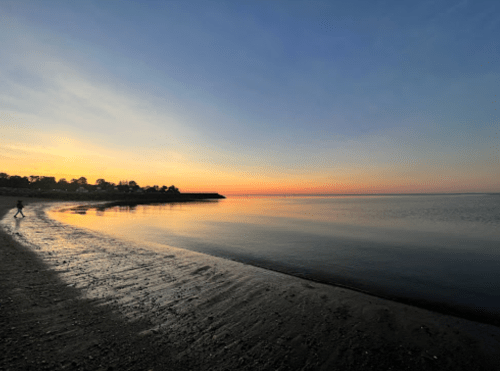 A serene beach at sunset, with calm waters and a person walking along the shore.