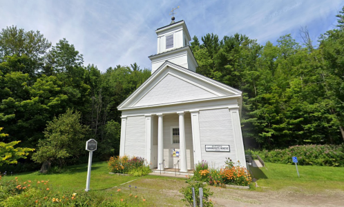 A white church building surrounded by greenery and flowers, with a sign and a clear blue sky above.