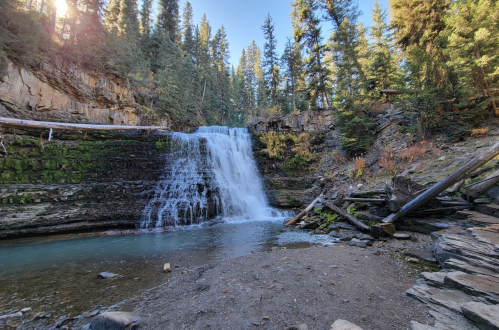 A serene waterfall cascades over rocky cliffs, surrounded by lush green trees and a clear blue sky.