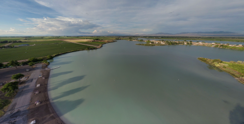 Aerial view of a calm lake surrounded by green fields and distant mountains under a cloudy sky.