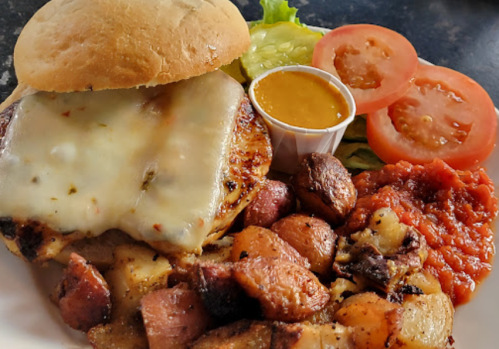 A grilled chicken sandwich with melted cheese, served with roasted potatoes, lettuce, tomato slices, and dipping sauce.