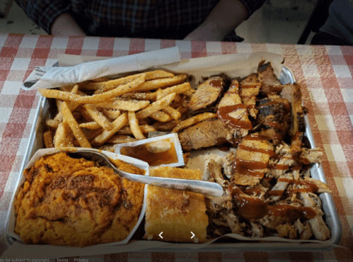 A tray of barbecue with sliced meats, fries, cornbread, and sauces on a checkered tablecloth.