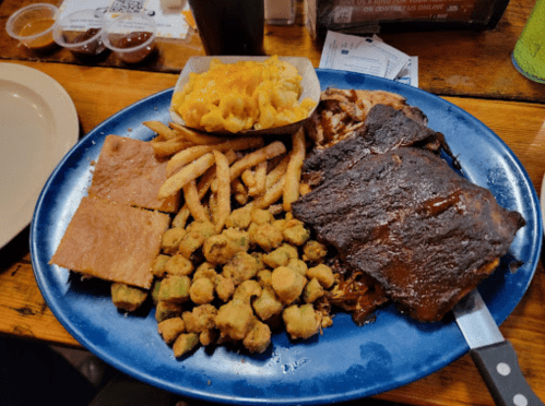 A blue plate filled with ribs, fries, cornbread, mac and cheese, and fried okra, set on a wooden table.