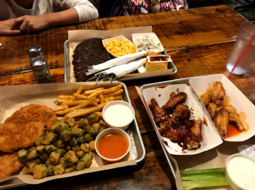 A table with two trays of food: fried chicken, okra, fries, ribs, macaroni, and wings with dipping sauces.
