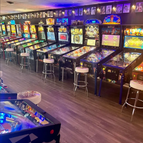 A row of colorful pinball machines in a game room, with stools and framed photos on the wall above.