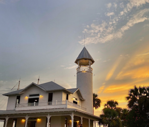 A white lighthouse-style building against a colorful sunset sky, surrounded by palm trees.