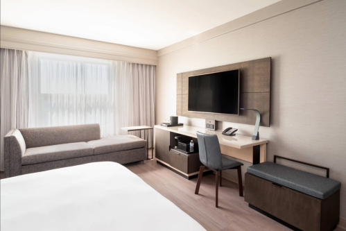 Modern hotel room featuring a sofa, desk, TV, and large window with sheer curtains.