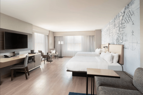 A modern hotel room featuring two beds, a large TV, a desk, and seating area with natural light from large windows.