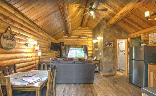 Cozy log cabin interior with wooden beams, a sofa, dining area, and kitchen appliances, creating a warm atmosphere.