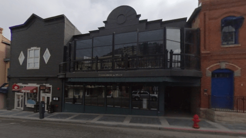 A modern two-story building with large glass windows, located on a city street, featuring a balcony and nearby shops.