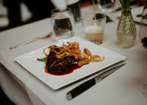 A plated steak with crispy onions, green beans, and roasted potatoes, served on a white tablecloth with drinks in the background.