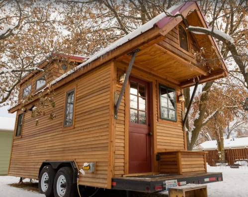 A cozy wooden tiny house on wheels, surrounded by snow-covered trees, featuring a sloped roof and front porch.