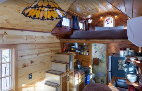 Cozy tiny house interior with wooden walls, a lofted bed, kitchen area, and decorative stained glass windows.
