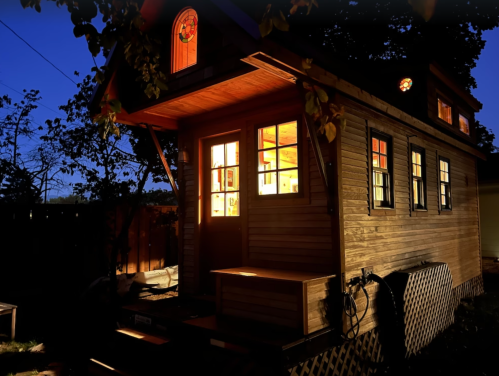 A cozy tiny house illuminated at night, with warm light glowing from its windows and surrounded by trees.