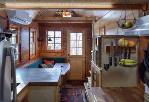 Cozy wooden interior of a tiny home with a kitchen, dining area, and natural light from windows and door.