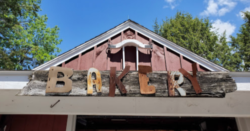 Sign reading "BAKERY" made of various materials, mounted on a rustic building with trees and blue sky in the background.