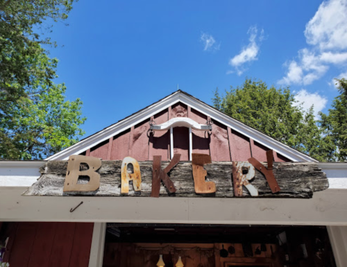 A rustic bakery sign made of weathered wood and metal letters against a blue sky with fluffy clouds.