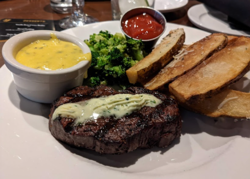 A plate featuring a grilled steak with herb butter, broccoli, potato wedges, and a side of cheese sauce and ketchup.