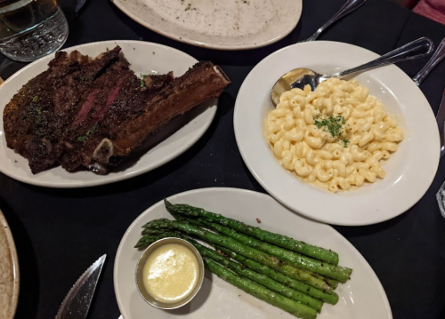A plate with a large ribeye steak, macaroni and cheese, and grilled asparagus with a small cup of sauce.