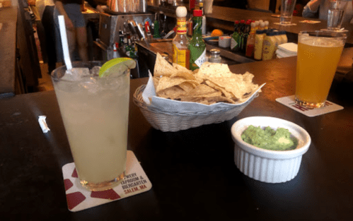 A refreshing drink with a lime, a basket of tortilla chips, and a small bowl of guacamole on a bar counter.