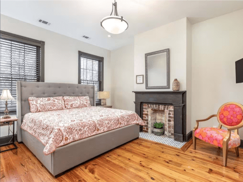 Cozy bedroom with a gray bed, patterned bedding, a fireplace, and a colorful chair, featuring wooden floors and natural light.