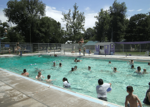A busy outdoor swimming pool with people swimming, playing, and lounging under a sunny sky. Trees in the background.