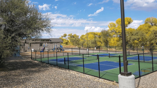 A tennis court with blue surfaces, surrounded by trees and a playground in the background under a partly cloudy sky.