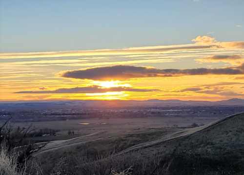 Sunset over rolling hills, with vibrant orange and yellow hues in the sky and a tranquil landscape below.
