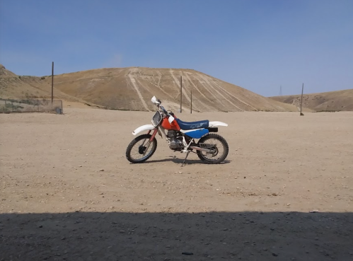 A dirt bike parked on a sandy terrain with a hilly background under a clear blue sky.
