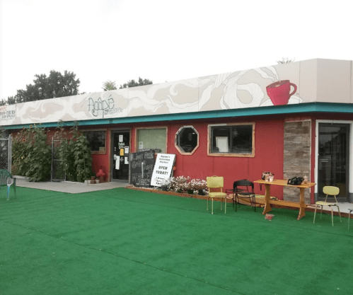 A colorful café building with a sign reading "Open Today," surrounded by greenery and outdoor seating on green turf.