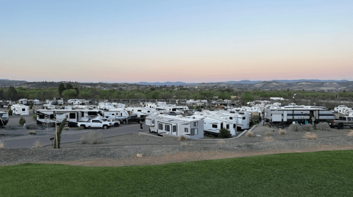 A panoramic view of a large RV park with numerous trailers and trucks, set against a backdrop of mountains at dusk.