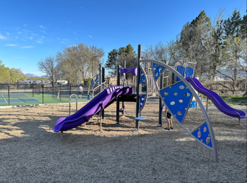 Colorful playground with purple slides and climbing structures, surrounded by trees and a tennis court in the background.