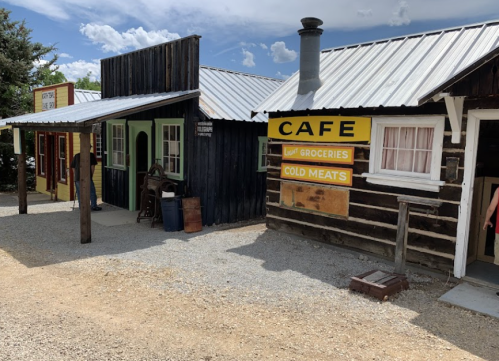 A rustic building with a café sign, featuring wooden structures and a gravel path in a small town setting.