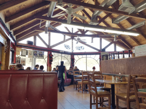 Interior of a rustic restaurant with wooden beams, tables, and patrons dining near large windows.