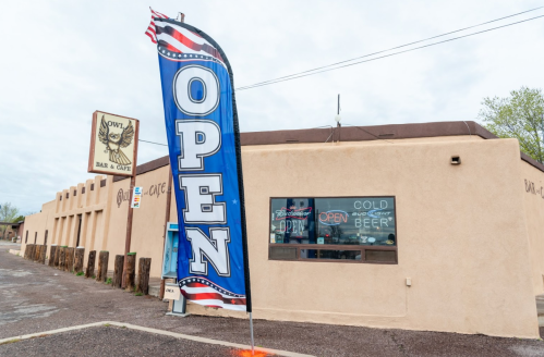 A bar and café with a large "OPEN" flag outside, featuring a rustic exterior and a sign for cold beer.