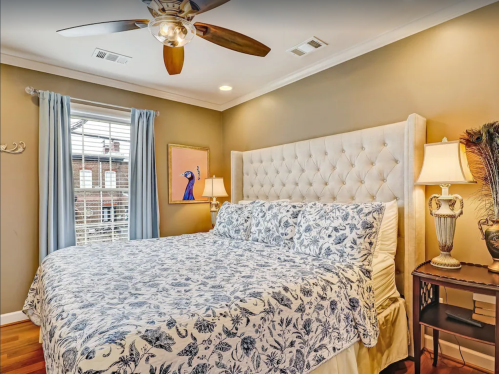 Cozy bedroom with a tufted headboard, floral bedding, and a ceiling fan, featuring soft lighting and a window view.