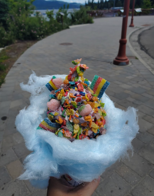 A colorful dessert topped with candy and cereal, served on a bed of blue cotton candy, held in a hand.