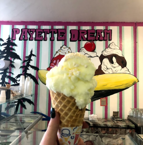 A hand holds a cone of yellow ice cream in front of a colorful mural featuring ice cream sundaes.